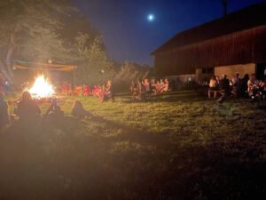 Abendlicht im Feuerschein. Teilnehmer des Festes sitzen um das Sonnwendfeuer im Hintergrund leuchtet der Mond